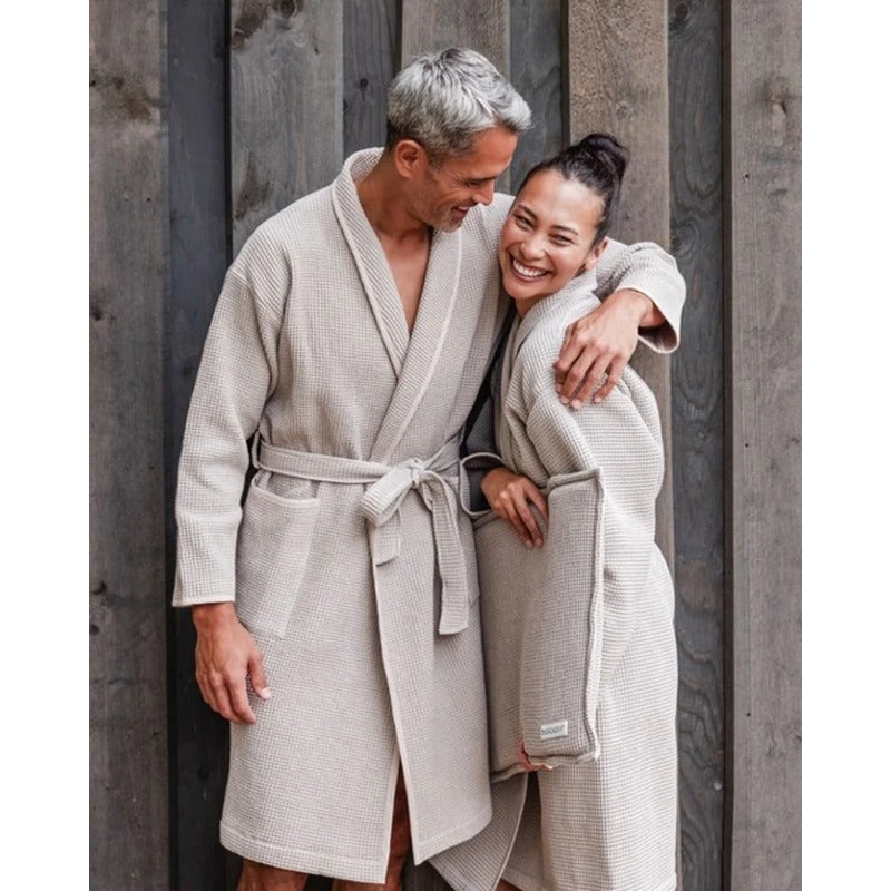 Ein Paar in Badesofa Bademänteln aus Bio-Baumwolle lehnt lachend aneinander vor einer Holzfassade.