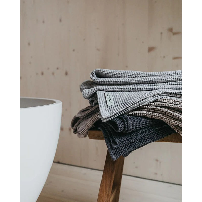 Gestapelte Badesofa Saunatücher aus Waffelpiqué in verschiedenen Naturtönen auf einer Holzbank neben einer Badewanne.